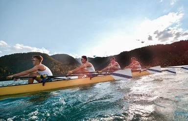 Vier Männer in sportlicher Kleidung rudern in einem Ruderboot auf einem See.