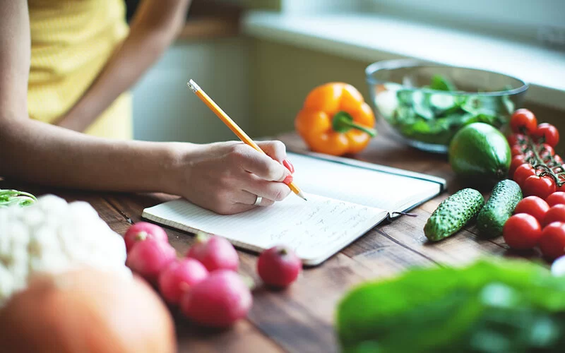 Eine Frau sitzt am Essenstisch mit Lebensmitteln und schreibt ein Ernährungstagebuch.