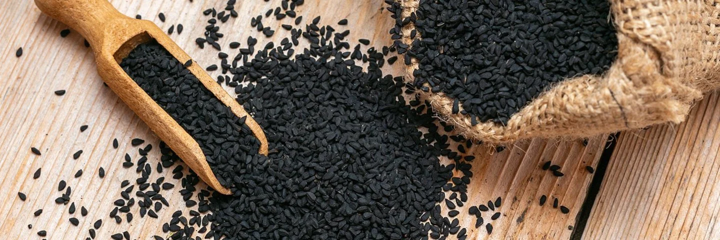 Samen von Schwarzkümmel liegen auf einem Tisch verstreut neben einem gefüllten Jutesack und einer kleinen Mehlschaufel aus Holz.