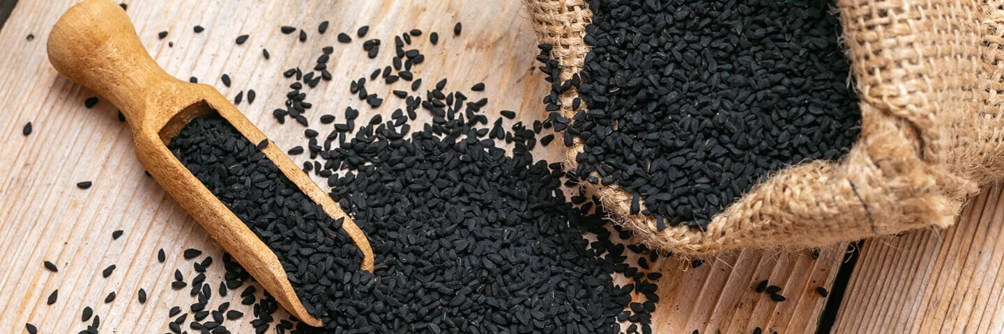 Samen von Schwarzkümmel liegen auf einem Tisch verstreut neben einem gefüllten Jutesack und einer kleinen Mehlschaufel aus Holz.