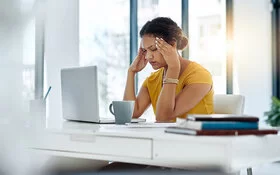 Eine Frau mit dunklen, hochgesteckten Haaren und gelbem Shirt sitzt in einem Büro, vor sich Laptop und Kaffeetasse. Die Frau hat die Augen geschlossen und fasst sich mit den Fingerspitzen an die Schläfen.