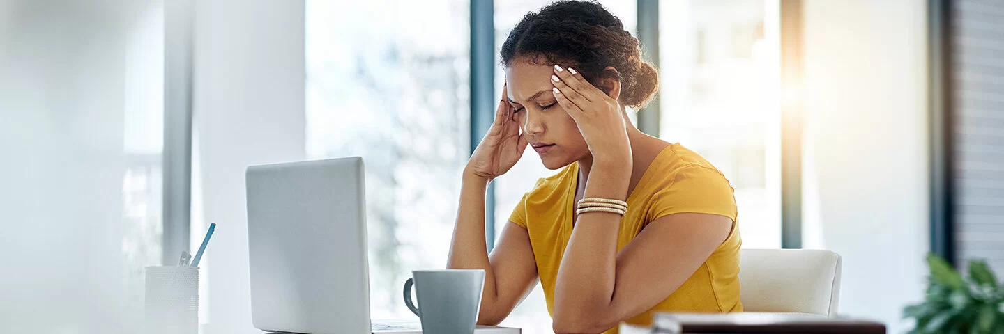 Eine Frau mit dunklen, hochgesteckten Haaren und gelbem Shirt sitzt in einem Büro, vor sich Laptop und Kaffeetasse. Die Frau hat die Augen geschlossen und fasst sich mit den Fingerspitzen an die Schläfen.