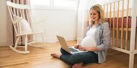 Eine schwangere Frau sitzt im Kinderzimmer auf dem Boden und lehnt sich mit dem Rücken an einem Kinderbett an. Auf ihrem Schoss hat sie einen Laptop und blickt auf den Bildschirm.