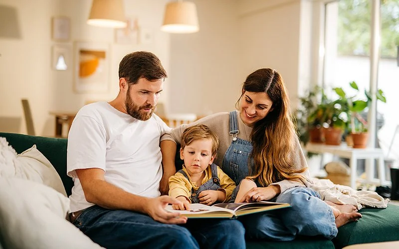 Auf einem Sofa im Wohnzimmer sitzen ein Mann und eine Frau mit einem Kind zwischen sich. Sie betrachten gemeinsam ein Buch.