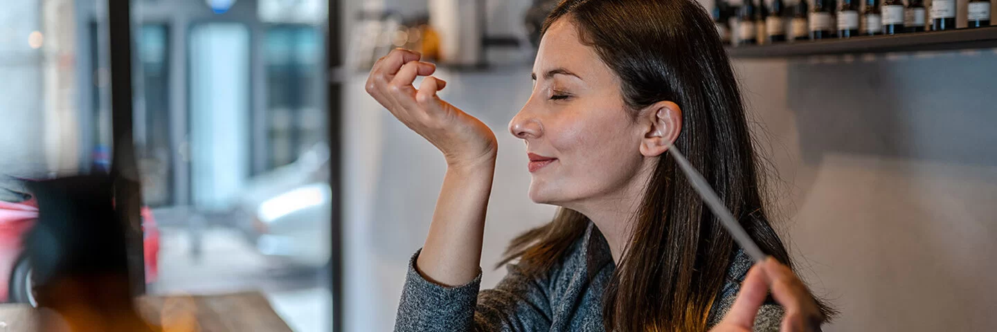Eine junge Frau hält in der linken Hand eine Pipette. Ihr rechtes Handgelenk hat sie an der Nase. Es wirkt, als ob der Geruch angenehm ist.