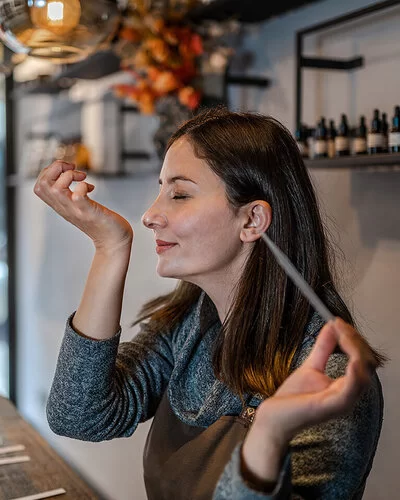 Eine junge Frau hält in der linken Hand eine Pipette. Ihr rechtes Handgelenk hat sie an der Nase. Es wirkt, als ob der Geruch angenehm ist.