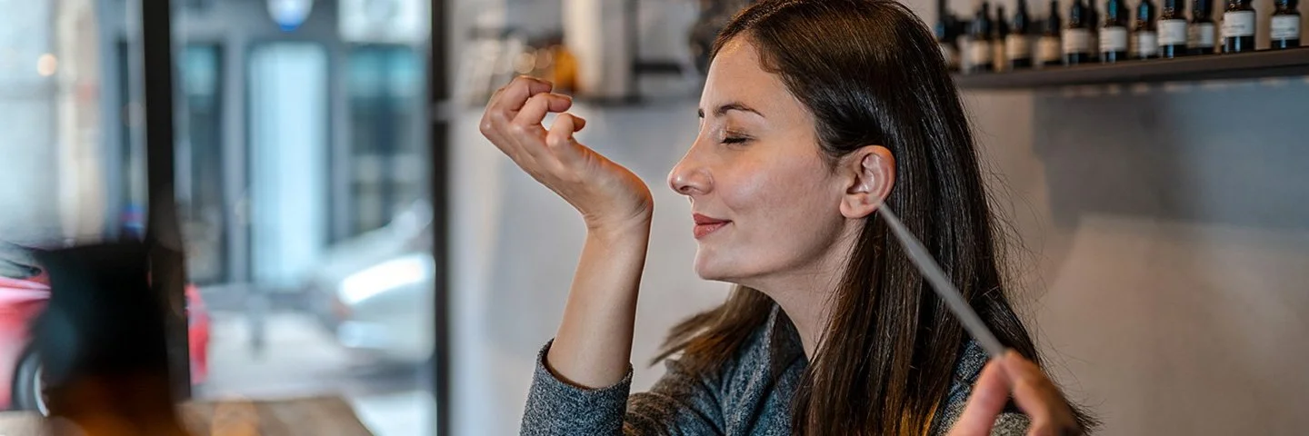 Eine junge Frau hält in der linken Hand eine Pipette. Ihr rechtes Handgelenk hat sie an der Nase. Es wirkt, als ob der Geruch angenehm ist.