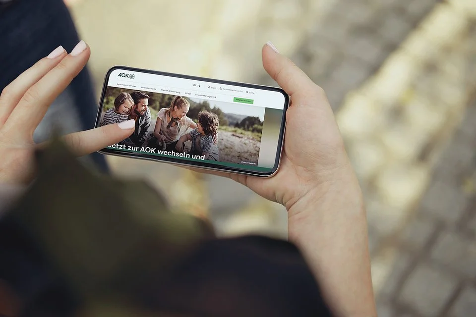 Eine nicht erkennbare Person hält ein Smartphone in den Händen, auf dem Display ist die Internetseite der AOk zu sehen.