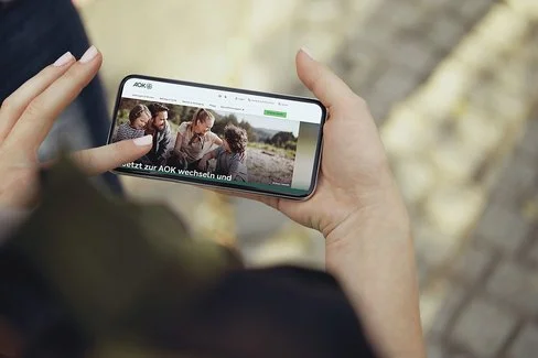 Eine nicht erkennbare Person hält ein Smartphone in den Händen, auf dem Display ist die Internetseite der AOk zu sehen.