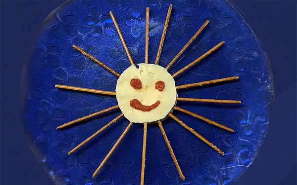 Auf einem blauen Teller liegt eine runde Knäckebrotscheibe mit Käse belegt. Ein Smiley aus Tomatenmark und Sonnenstrahlen aus Salzstangen machen den Snack zu einer essbaren Sonne.