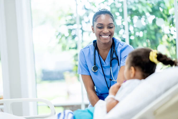 Eine Frau in blauer Arbeitskleidung lächelt ein Mädchen an, das in einem Krankenhausbett liegt.