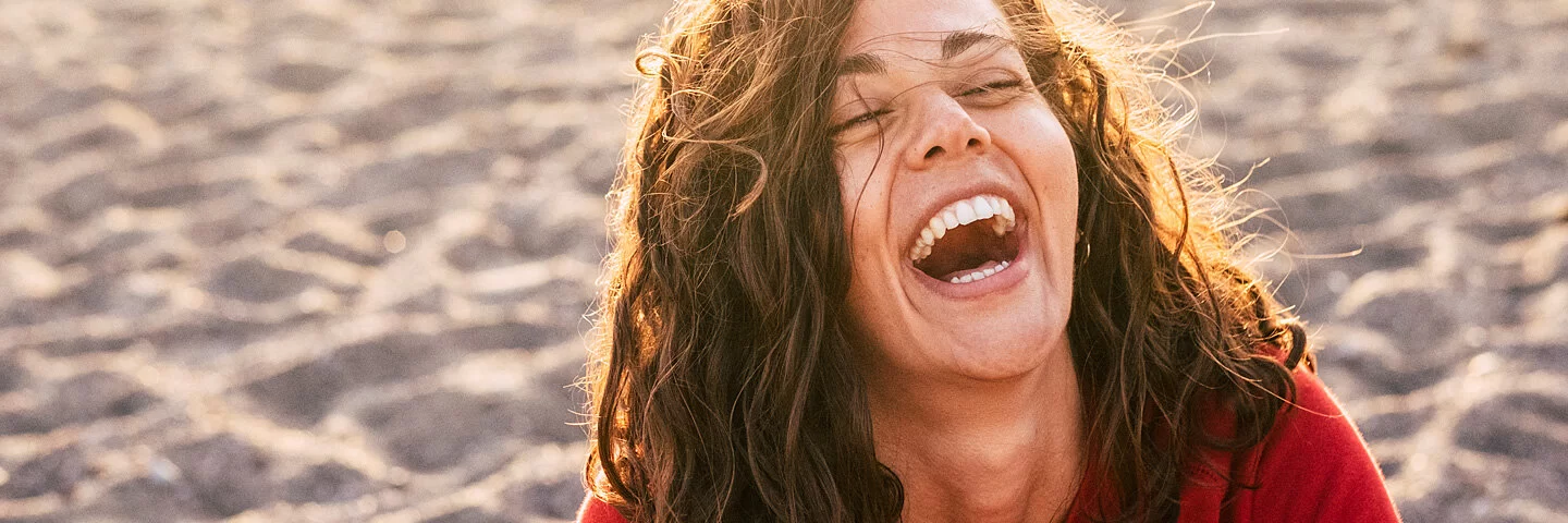 Eine Frau am Strand lacht herzhaft im Sonnenuntergang.