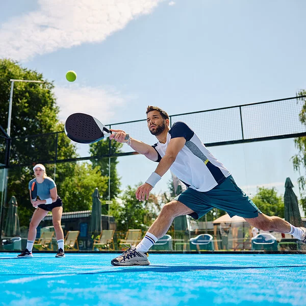 Ein junger Mann macht auf einem Padel-Feld einen Ausfallschritt, um den Ball noch zu erreichen. Er spielt ihn mit dem Schläger.