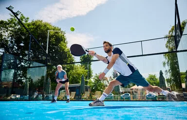 Ein junger Mann macht auf einem Padel-Feld einen Ausfallschritt, um den Ball noch zu erreichen. Er spielt ihn mit dem Schläger.