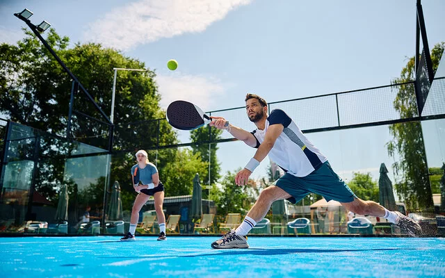 Ein junger Mann macht auf einem Padel-Feld einen Ausfallschritt, um den Ball noch zu erreichen. Er spielt ihn mit dem Schläger.