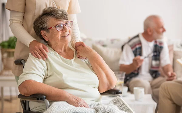 Eine lächelnde Seniorin sitzt in einem Rollstuhl in einem Pflegeheim und eine Pflegerin steht hinter ihr.