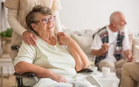 Eine lächelnde Seniorin sitzt in einem Rollstuhl in einem Pflegeheim und eine Pflegerin steht hinter ihr.