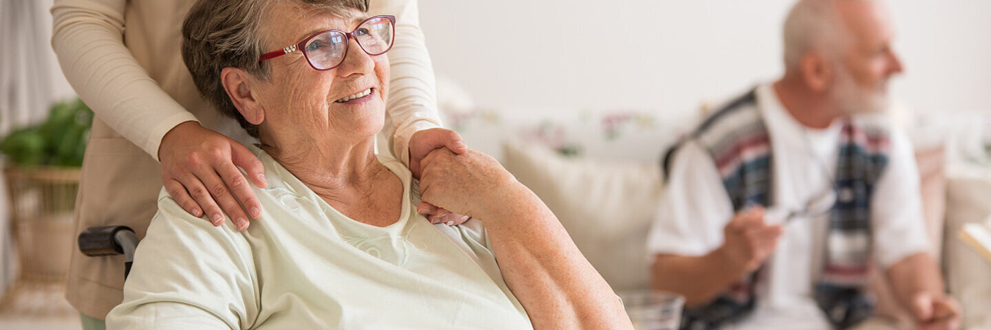 Eine lächelnde Seniorin sitzt in einem Rollstuhl in einem Pflegeheim und eine Pflegerin steht hinter ihr.