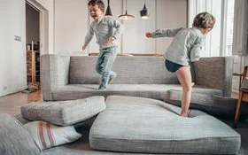 Zwei Jungen toben auf einem grauen Sofa und auf dem Boden davor liegenden Sofakissen in einem hellen Wohnzimmer.