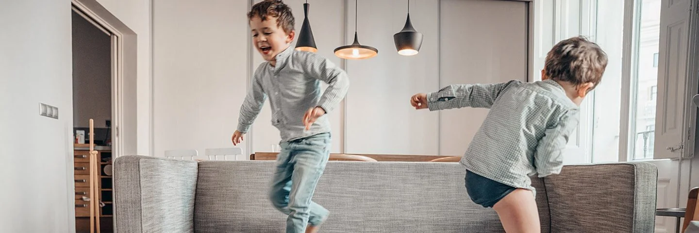 Zwei Jungen toben auf einem grauen Sofa und auf dem Boden davor liegenden Sofakissen in einem hellen Wohnzimmer.