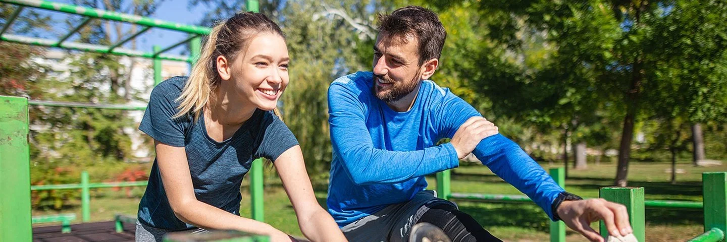 An einem sonnigen Tag machen eine junge Frau und ein junger Mann in Sportkleidung gemeinsam Dehnübungen für die Beine in einer Outdoor-Fitnessanlage. Beide lächeln, während der Mann die Frau anschaut.