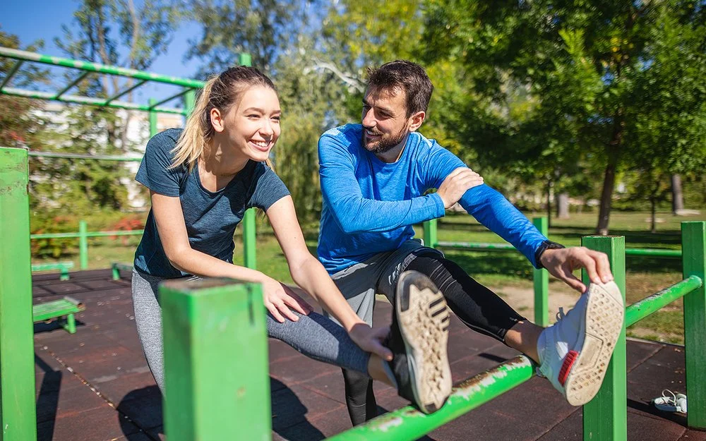 An einem sonnigen Tag machen eine junge Frau und ein junger Mann in Sportkleidung gemeinsam Dehnübungen für die Beine in einer Outdoor-Fitnessanlage. Beide lächeln, während der Mann die Frau anschaut.