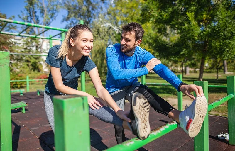 An einem sonnigen Tag machen eine junge Frau und ein junger Mann in Sportkleidung gemeinsam Dehnübungen für die Beine in einer Outdoor-Fitnessanlage. Beide lächeln, während der Mann die Frau anschaut.