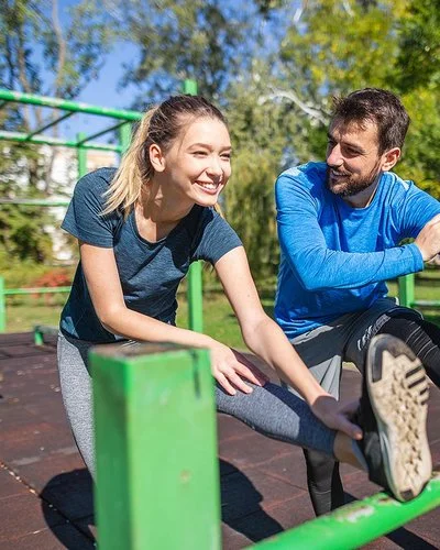 An einem sonnigen Tag machen eine junge Frau und ein junger Mann in Sportkleidung gemeinsam Dehnübungen für die Beine in einer Outdoor-Fitnessanlage. Beide lächeln, während der Mann die Frau anschaut.