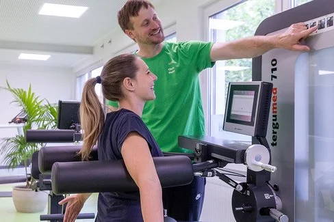 Eine Frau sitzt an einem Rückenübungsgerät im Fitnessstudio, ihr Trainer zeigt auf die Gewichtseinstellung am Gerät