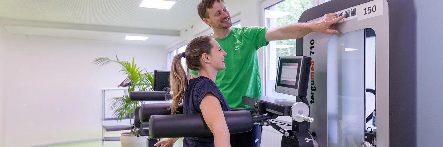 Eine Frau sitzt an einem Rückenübungsgerät im Fitnessstudio, ihr Trainer zeigt auf die Gewichtseinstellung am Gerät