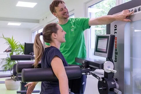 Eine Frau sitzt an einem Rückenübungsgerät im Fitnessstudio, ihr Trainer zeigt auf die Gewichtseinstellung am Gerät