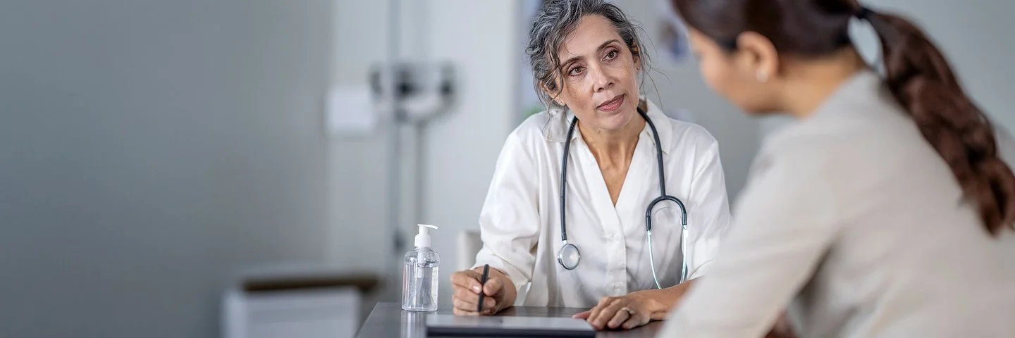 Ärztin mit Stethoskop sitzt an einem Tisch und spricht mit einer Patientin im Vordergrund.