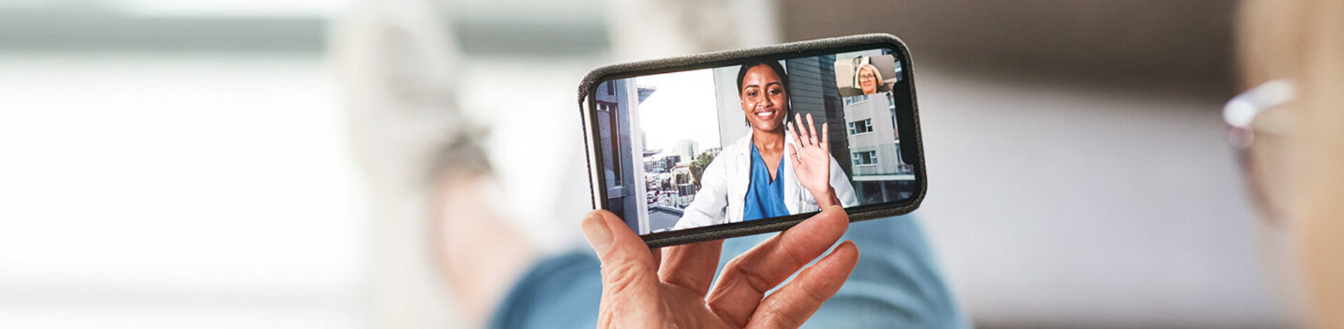 Eine Frau hält ein Handy in der Hand, auf dessen Bildschirm eine Frau in einem weißen Kittel zu sehen ist.