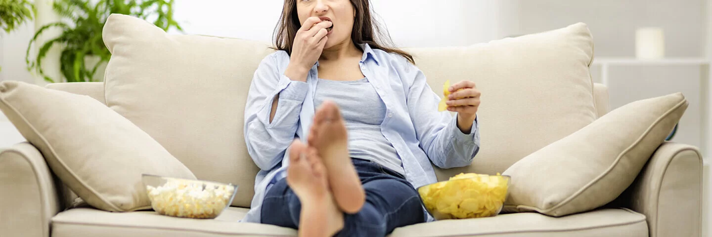 Frau sitzt auf einem Sofa zwischen einer Schüssel Chips und einer Schüssel Popcorn – sie  leidet unter emotionalem Essverhalten.