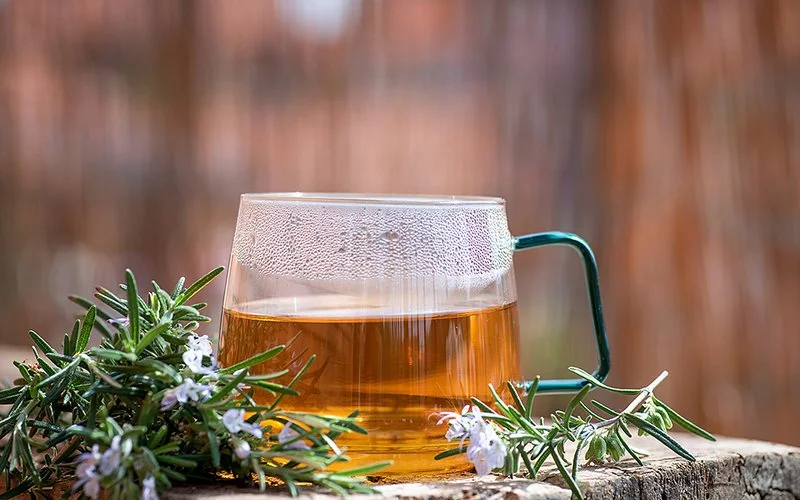 Ein Glas Rosmarintee steht auf einem hölzernem Untergrund, daneben liegen blühende Rosmarinzweige.