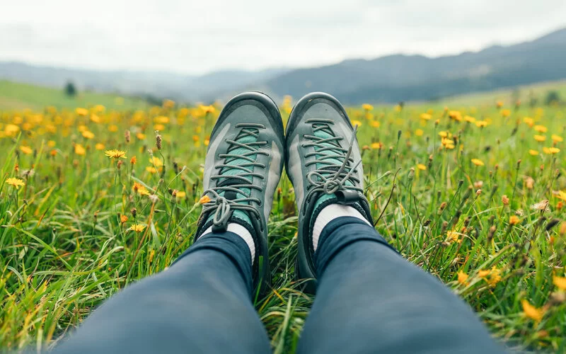 Die mit Wanderschuhen bekleideten Füße einer Pilgerin sind auf einer Blumenwiese zu sehen.