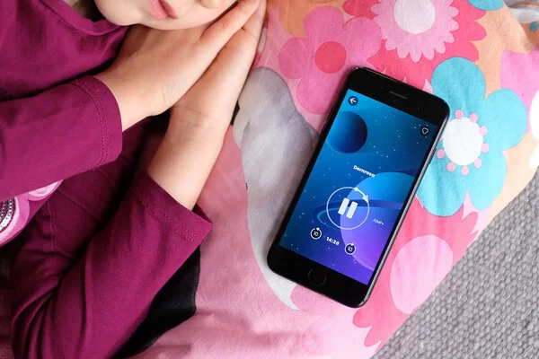 Ein nicht erkennbares Kind liegt schlafend auf einem Bett, im Fokus des Bildes ist ein Smartphone mit laufender Aumio-App zu sehen.