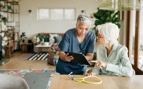 Eine weibliche Pflegekraft sitzt zusammen mit einer alten Frau in einem Wohnzimmer an einem Tisch. Die Pflegerin hält ein Tablet in den Händen und beide schauen auf den Bildschirm.