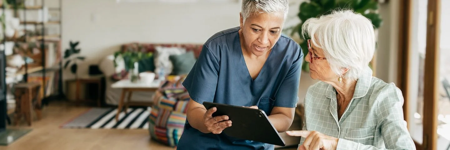 Eine weibliche Pflegekraft sitzt zusammen mit einer alten Frau in einem Wohnzimmer an einem Tisch. Die Pflegerin hält ein Tablet in den Händen und beide schauen auf den Bildschirm.
