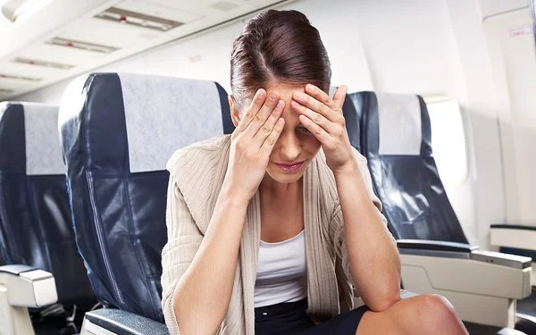 Eine Frau sitzt mit schmerzverzerrtem oder erschöpftem Gesichtsausdruck in einem Flugzeugsitz und hält sich die Hände vor die Stirn.