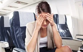 Eine Frau sitzt mit schmerzverzerrtem oder erschöpftem Gesichtsausdruck in einem Flugzeugsitz und hält sich die Hände vor die Stirn.