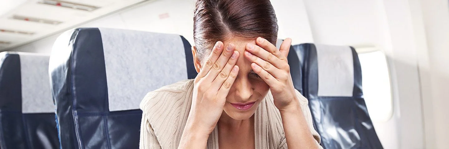 Eine Frau sitzt mit schmerzverzerrtem oder erschöpftem Gesichtsausdruck in einem Flugzeugsitz und hält sich die Hände vor die Stirn.
