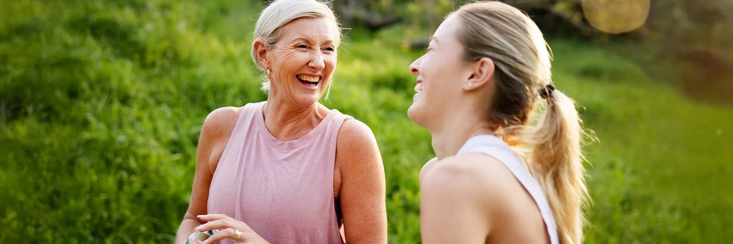 Eine mittelalte Frau und ihre erwachsene Tochter in Sportkleidung lachen beherzt in grüner Umgebung.