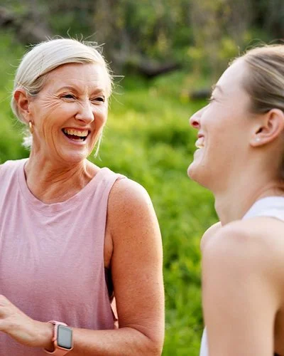 Eine mittelalte Frau und ihre erwachsene Tochter in Sportkleidung lachen beherzt in grüner Umgebung.
