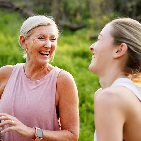 Eine mittelalte Frau und ihre erwachsene Tochter in Sportkleidung lachen beherzt in grüner Umgebung.