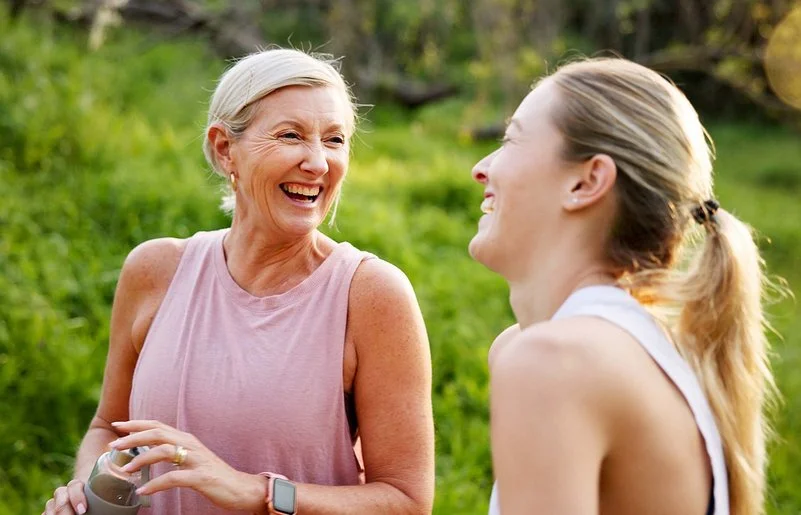 Eine mittelalte Frau und ihre erwachsene Tochter in Sportkleidung lachen beherzt in grüner Umgebung.