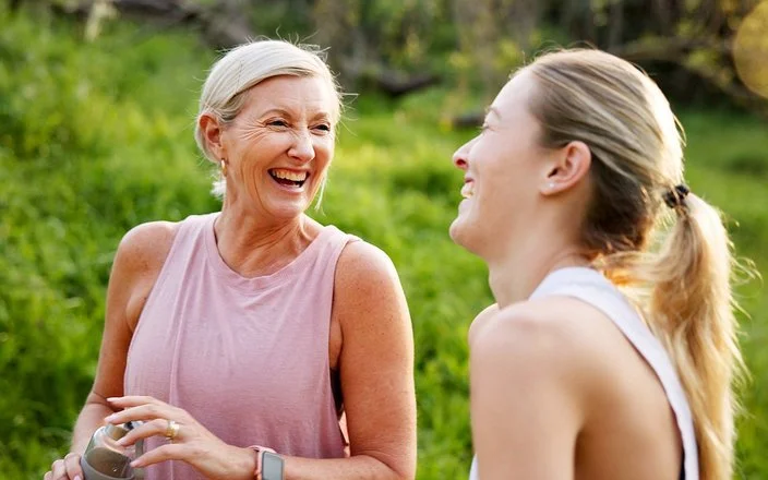 Eine mittelalte Frau und ihre erwachsene Tochter in Sportkleidung lachen beherzt in grüner Umgebung.