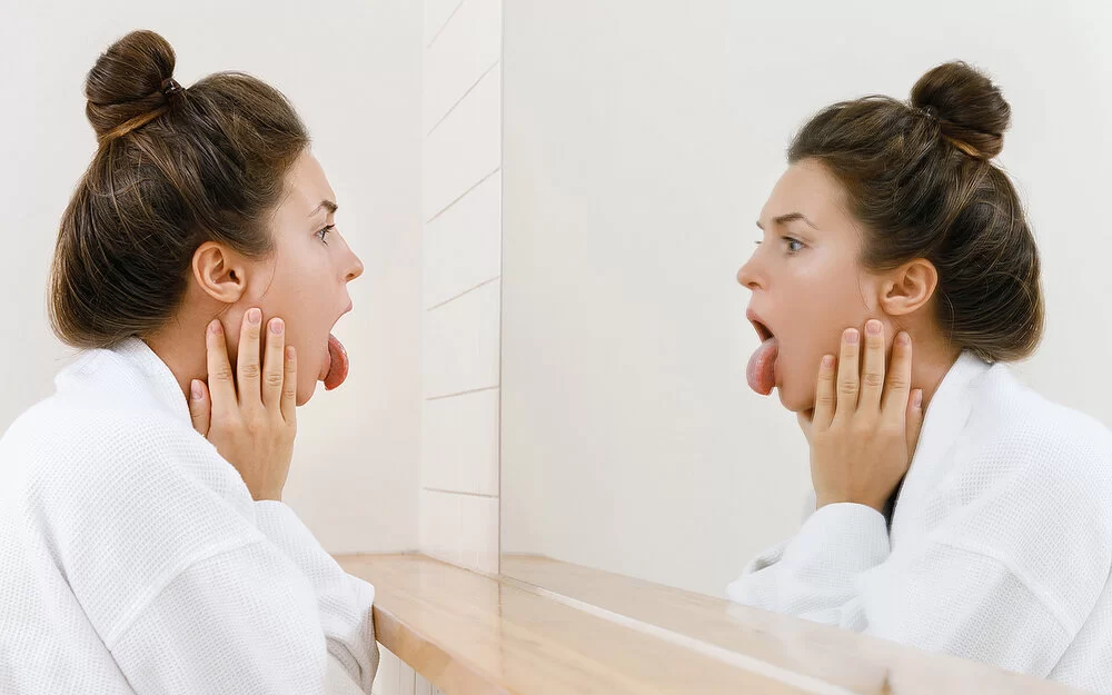 Eine junge Frau mit Dutt schaut vor dem Badezimmerspiegel auf ihre Zunge und untersucht sich auf Mundsoor.