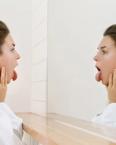 Eine junge Frau mit Dutt schaut vor dem Badezimmerspiegel auf ihre Zunge und untersucht sich auf Mundsoor.