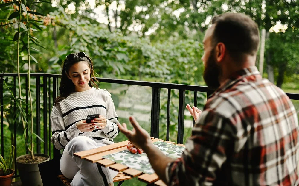 Auf einem Balkon zum Garten sitzen sich eine junge Frau und ein junger Mann an einem Tisch gegenüber. Die Frau liest lächelnd auf ihrem Smartphone. Der Mann hebt gestikulierend beide Hände.